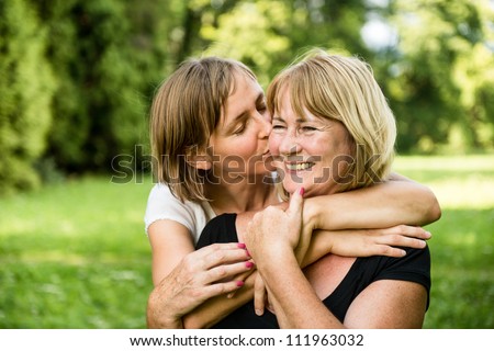Adult daughter kissing her smiling happy senior mother - outdoor in nature