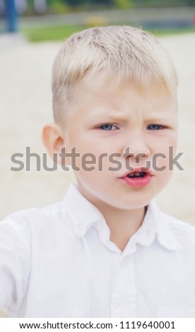 Portrait of a little displeased and dissatisfied boy with golden blonde hair in sunny summer day on green blurred background.