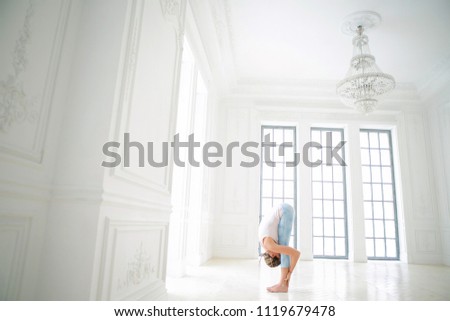 Young attractive woman practicing yoga. Working out wearing sportswear, white T-shirt and blue pants. Standing forward bend, head to knees exercise, uttanasana pose.