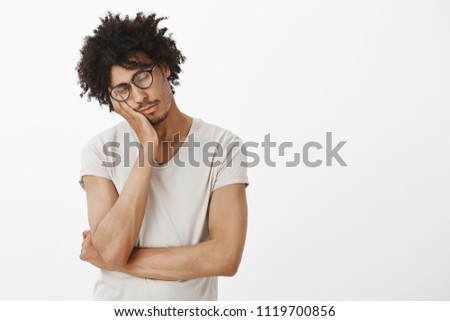 Indoor shot of cute sleepy male hispanic model with curly hair in glasses and grey t-shirt, leaning on palm, closing eyes, sleeping, feeling tired and exhausted, spending sleepless night at work