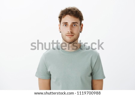 Close-up shot of ordinary fair-haired caucasian male with bristle in earrings, staring carefree at camera being indifferent and calm, listening to boss while standing against gray background Royalty-Free Stock Photo #1119700988
