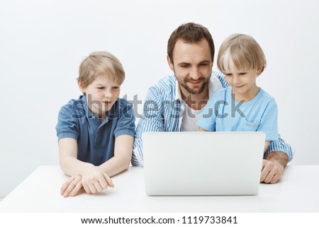 Indoor shot of positive happy blond child with father and brother sitting at table, looking at laptop screen and smiling broadly, having fun while watching cartoons or talking with mom via gadget
