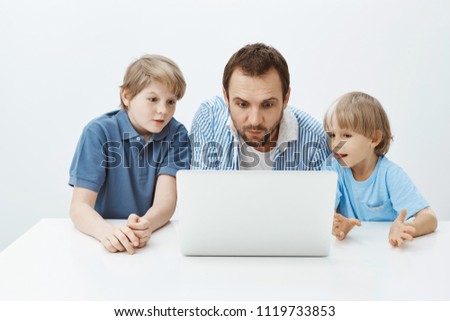 Father watching cartoons with sons. Portrait of enterteined attractive father with young boys sitting at table and looking at laptop screen with interest and curiousity, spending time with family