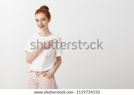 Intrigued charming stylish woman with red hair in bun hairstyle, pointing at upper right corner and smiling broadly at camera, asking question, being interested in price of cute t-shirt in shop Royalty-Free Stock Photo #1119734150