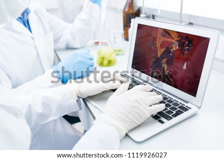 Crop view of microbiologists sitting at table testing samples of green vegetables and looking at computer model of food DNA on laptop screen 
