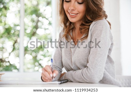 Cropped photo of cute cheerful young lady sitting in classroom writing notes.
