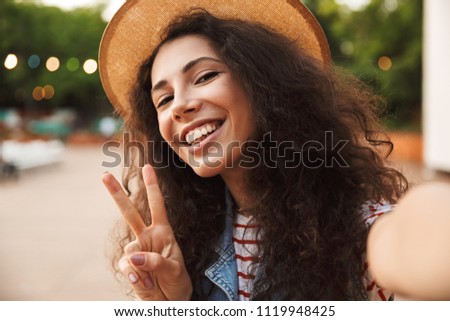 Photo closeup of beautiful teenage woman 18-20 wearing straw hat showing peace sign and taking selfie while walking outdoor
