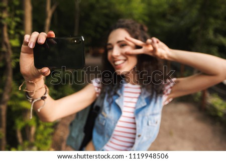 Blurry photo of brunette teenage woman 18-20 with backpack showing peace sign and taking selfie photo on smartphone while walking in green park