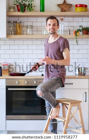 Portrait of brunet man with frying pan and phone in his hands in kitchen