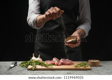 The man sprinkles the steak fillets of the minion with pepper salt. The chef works in the open kitchen of the restaurant. Fresh meat, garlic and rosemary on a wooden board. Black background