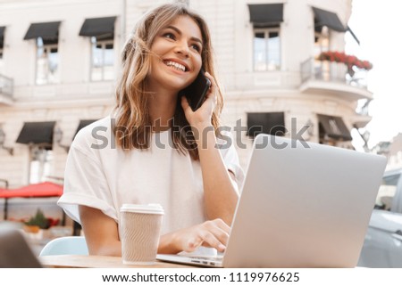 Portrait of happy young woman sitting at table in cafe outdoor on summer day and and having mobile call on smartphone while using silver laptop