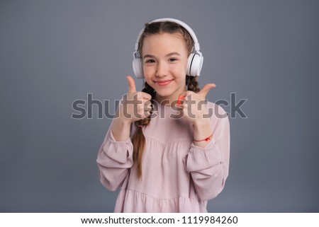 Great song. Adorable teenage girl listening to music in big headphones and showing thumbs up while posing isolated on a blue-grey background Royalty-Free Stock Photo #1119984260