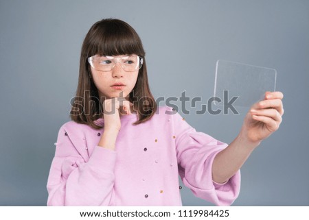 Cutting-edge technology. Charming teenage girl in smart glasses using a transparent smartphone while posing isolated on a blue-grey background