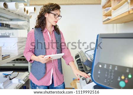 Adult woman in casual outfit holding tablet and setting printer while working in printing office. 