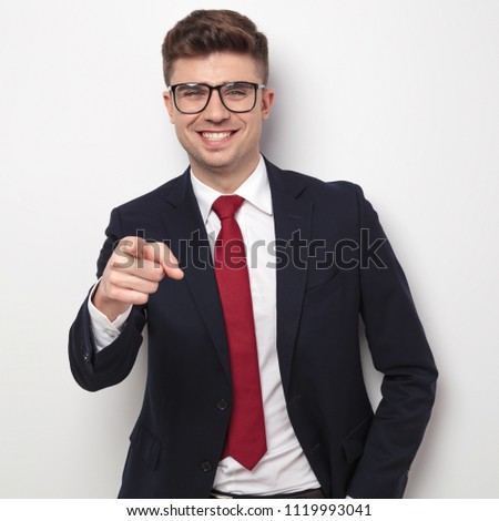 smiling businessman with glasses and navy suit points finger while standing on light grey background, portrait picture