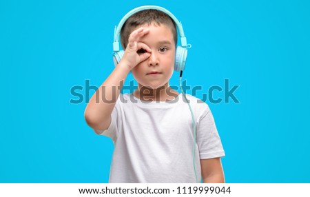 Dark haired little child listening music with headphones with happy face smiling doing ok sign with hand on eye looking through fingers
