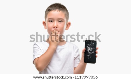 Dark haired little child holding broken smartphone cover mouth with hand shocked with shame for mistake, expression of fear, scared in silence, secret concept