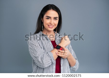 Useful device. Beautiful dark-haired woman demonstrating her smart watch and smiling pleasantly while posing isolated on a grey-blue background