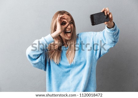 Smiling young blonde girl taking a selfie and showing ok gesture isolated over gray background