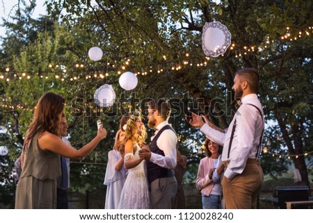 Guests with smartphones taking photo of bride and groom at wedding reception outside.