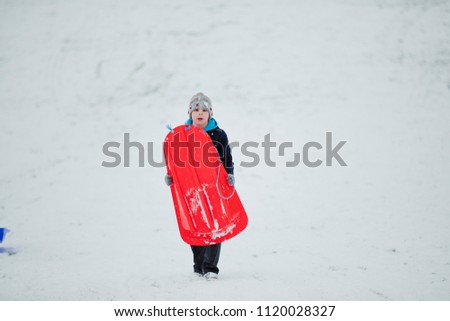 Little boy is walking toward the camera in a public park while it is snowing and is carrying a sled.