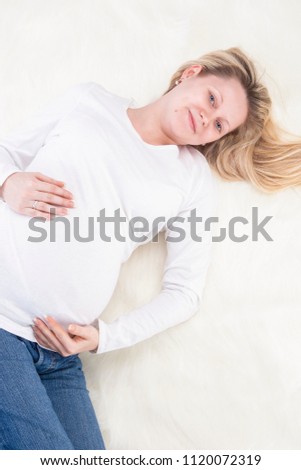 Family and Child-Bearing Concepts. Smiling Pregnant Caucasian Woman Laying on Fur Carpet In White Shirt and Jeans.Holding Her Abdomen. Vertical Image Composition