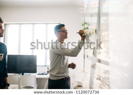 Man writing on board in the meeting room while his colleague looks on. Application developers in the board room discussing ideas and writing on board having adhesive post its on it.