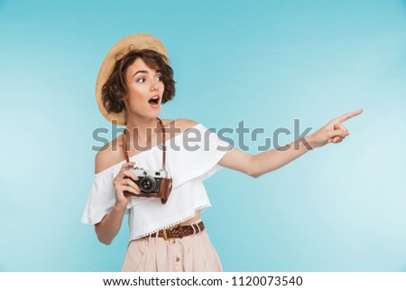 Portrait of an excited young woman in summer hat standing with photo camera and pointing away at copy space isolated over blue background
