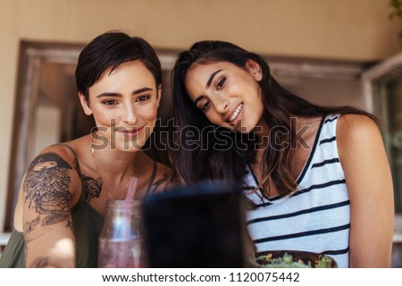 Two women sitting at a restaurant looking at a mobile phone. Women shooting selfie with food on the table at a restaurant.