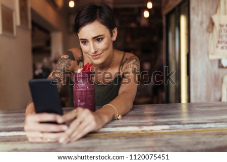 Woman taking a selfie with a smoothie placed in front of her using a mobile phone for her food blog. Food blogger shooting photos for her blog at home.