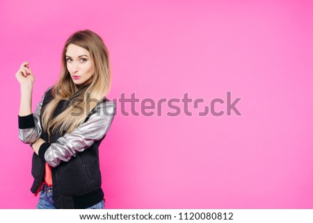 Stylish teen girl wearing in fashionable jeans and jacket posing and confident looking at camera. Blonde woman with perfect skin and natural make up standing with crossed hands in pink studio.