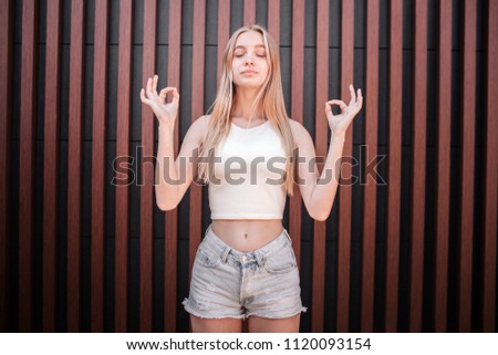 Light-haired girl is standing and meditating. She has her fingers in rounds. Girl is keeping her eyes closed. She is very calm and relaxed. Isolated on striped background.