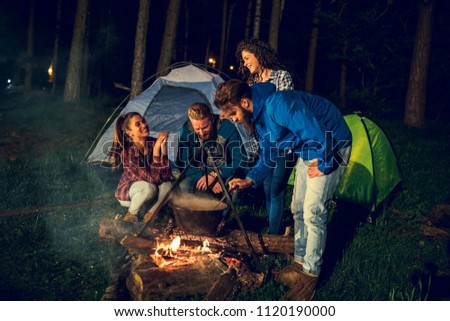 Group of friends cooking dinner over a campfire while camping in the woods.