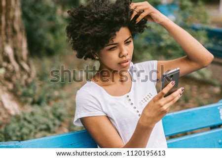 People, lifestyle. Portrait of attractive afro american woman sitting on bench outdoors in the park checking his news feed or messaging via social networks using smart phone using free wireless.