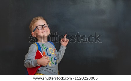 Back to school. Funny little boy in glasses pointing up on blackboard. Child from elementary school with book and bag. Education. Royalty-Free Stock Photo #1120228805