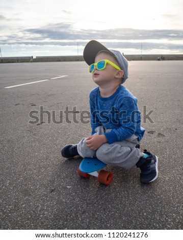 Street sports for children: A small boy in a cap and in sunglasses sat down on a blue skateboard to relax.