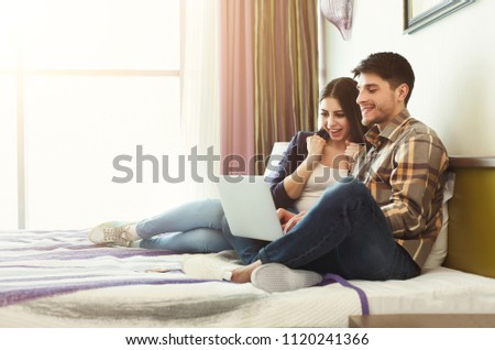Happy couple planning honeymoon trip on laptop while sitting on bed in hotel room