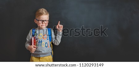Back to school. Funny little boy in glasses pointing up on blackboard. Child from elementary school with book and bag. Education.
