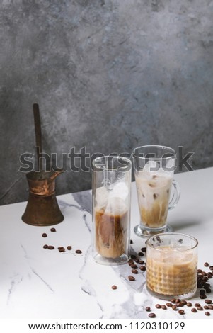 Iced coffee cocktail or frappe with ice cubes and cream served in three different glasses with vintage jezva and coffee beans around on white marble table with grey concrete wall at background.