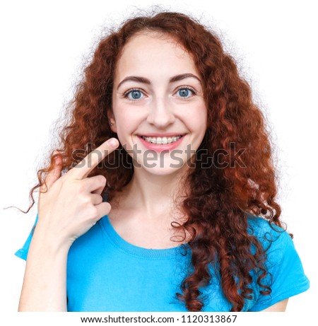 Girl with braces. Young european woman smiling in ceramic braces. Pointing a finger at her teeth. Dressed in a blue T-shirt. Happy smile. Girl with curly red hair Isolated on white background