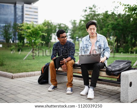 Two students from different countries live in a campus. Students discuss their project sitting on a bench in the Park. The photo illustrates student life, education at University or College.