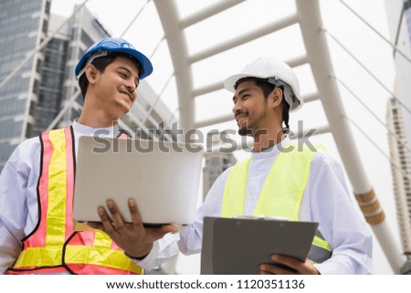 Two Handsome smile Burmese or Myanmar engineer manager and electrician using computer laptop to check construction project plan progress in Bangkok Modern city. Industrial concept.