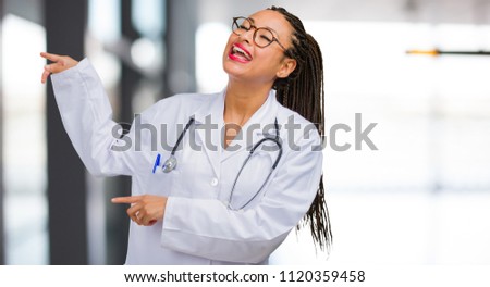 Portrait of a young black doctor woman pointing to the side, smiling surprised presenting something, natural and casual