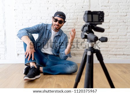 young attractive man in casual clothes hat beard hipster style with video camera on tripod against brick wall background in internet blog and blogger concept.