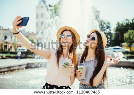 Hipster girls in sunglasses best friends drink cocktails doing self photo and laughing in city street