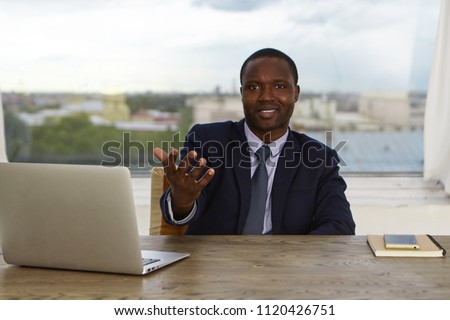 Portrait of positive Afro American male human resource manager in elegant suit sitting in his modern office in front of open laptop computer, reaching out hand to camera, greeting you at large company