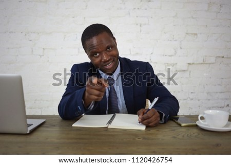 Horizontal shot of successful confident young African American chief executive officer dressed in elegant wear smiling and pointing index finger at camera, making notes in copybook. Body language
