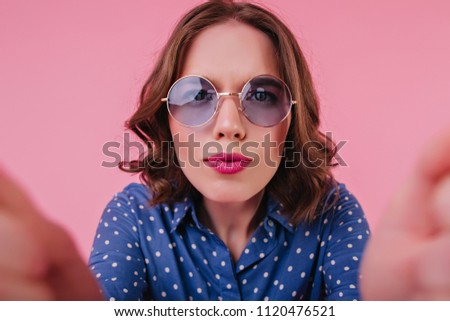 Funny young woman with bright makeup making selfie in studio. Indoor photo of good-looking female model in blue blouse taking picture of herself.
