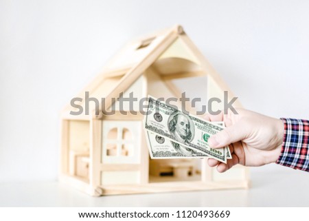 Hand giving one hundred us dollar banknotes. Wooden house model on background. Credit , debt or loan , real estate concept