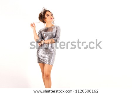 young girl in a silver shiny disco dress with curls and big crown is standing flirting with things on a white wall background